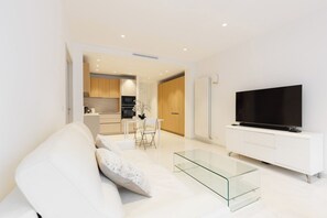 A modern living room with a sofa, a glass coffee table, a TV on a white cabinet, and a kitchen in the background. - Croisette : chic 2 chambres/ 2 salles de bains (Cannes)
