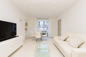 A modern living room with a flat-screen TV, a glass coffee table, a white sofa, and a dining area with a table and chairs. - Croisette : chic 2 chambres/ 2 salles de bains (Cannes)