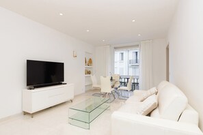 A modern living room with a flat-screen TV, a glass coffee table, a white sofa, and a dining area with chairs and a table. - Croisette : chic 2 chambres/ 2 salles de bains (Cannes)
