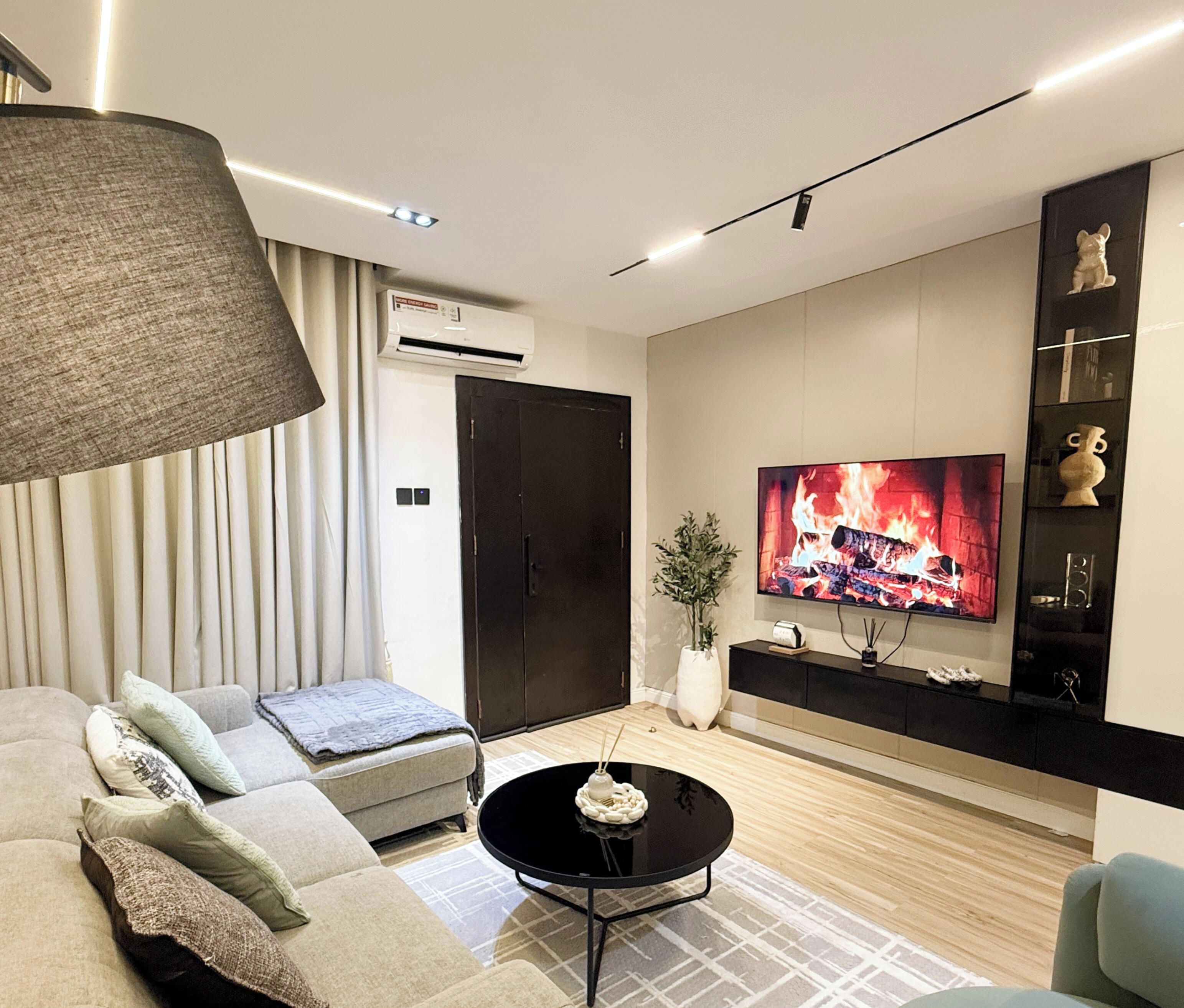 A modern living room with a sofa, a coffee table, a TV displaying a fireplace, and a wall-mounted shelf with decorative items.