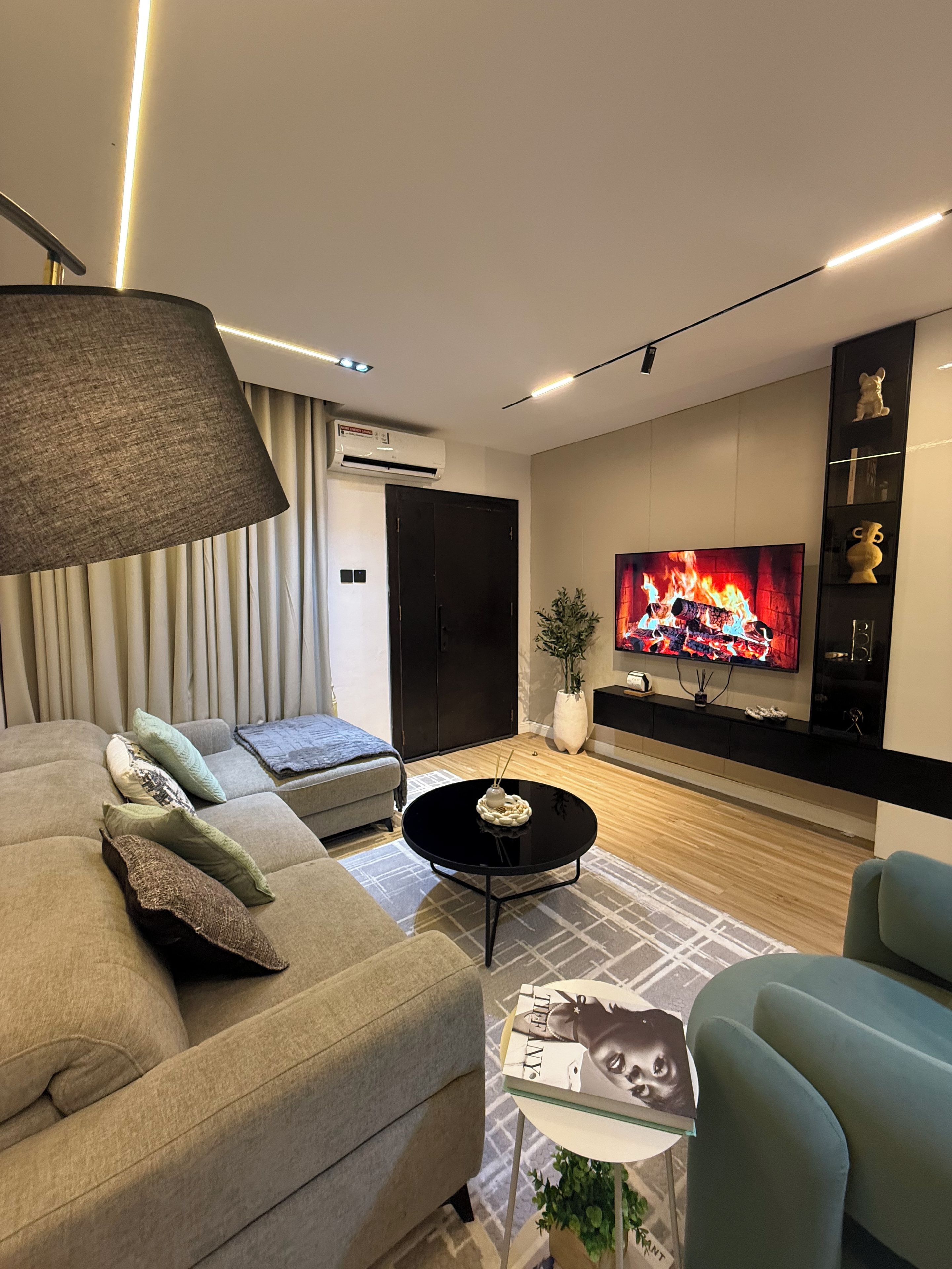 Ein modernes Wohnzimmer mit einer Couch, einem Couchtisch, einem Fernseher, der einen Kamin zeigt, und einer großen Lampe.