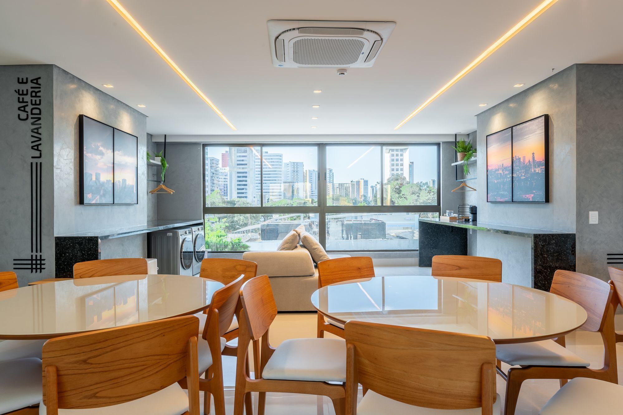 A modern interior with a central round table surrounded by wooden chairs, large windows offering a city view, and a visible laundry area.