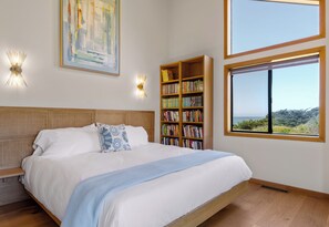 A bedroom with a large bed, a bookshelf, a painting, and a window with a view. - Oceanhaus, by Vinifera Homes (Sea Ranch)
