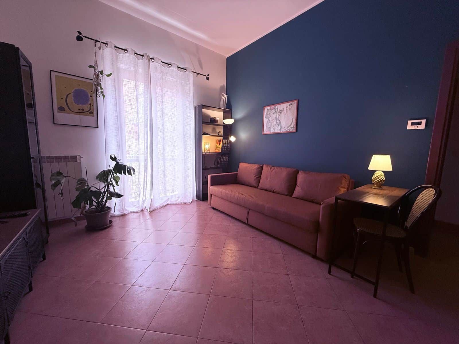 Ein Wohnzimmer mit einer Couch, einem Tisch mit einer Lampe, einem Topfpflanzen und einem Bild an der Wand.