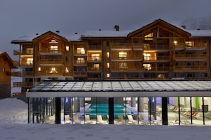 A multi-story wooden building with balconies, a glass-enclosed pool area, and snow-covered surroundings. - Résidence CGH les Chalets de Flambeau (Val-Cenis)