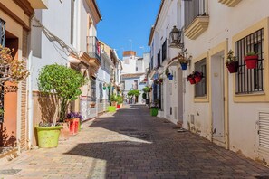 Un carrer pavimentat i estret flanquejat per edificis blancs, alguns amb balcons i plantes en testos.