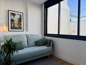 A light blue sofa with green cushions, a framed picture on the wall, and a window with a view of a building. - Coral8 - Top modern townhouse in Estepona old town (Estepona)