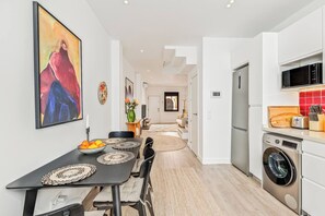 Une cuisine moderne équipée d’un lave-linge, un coin repas avec une table dressée pour deux personnes, et un salon en arrière-plan.