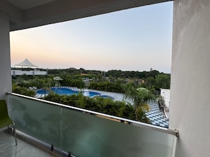 A balcony with a view of a swimming pool surrounded by greenery and a distant building.