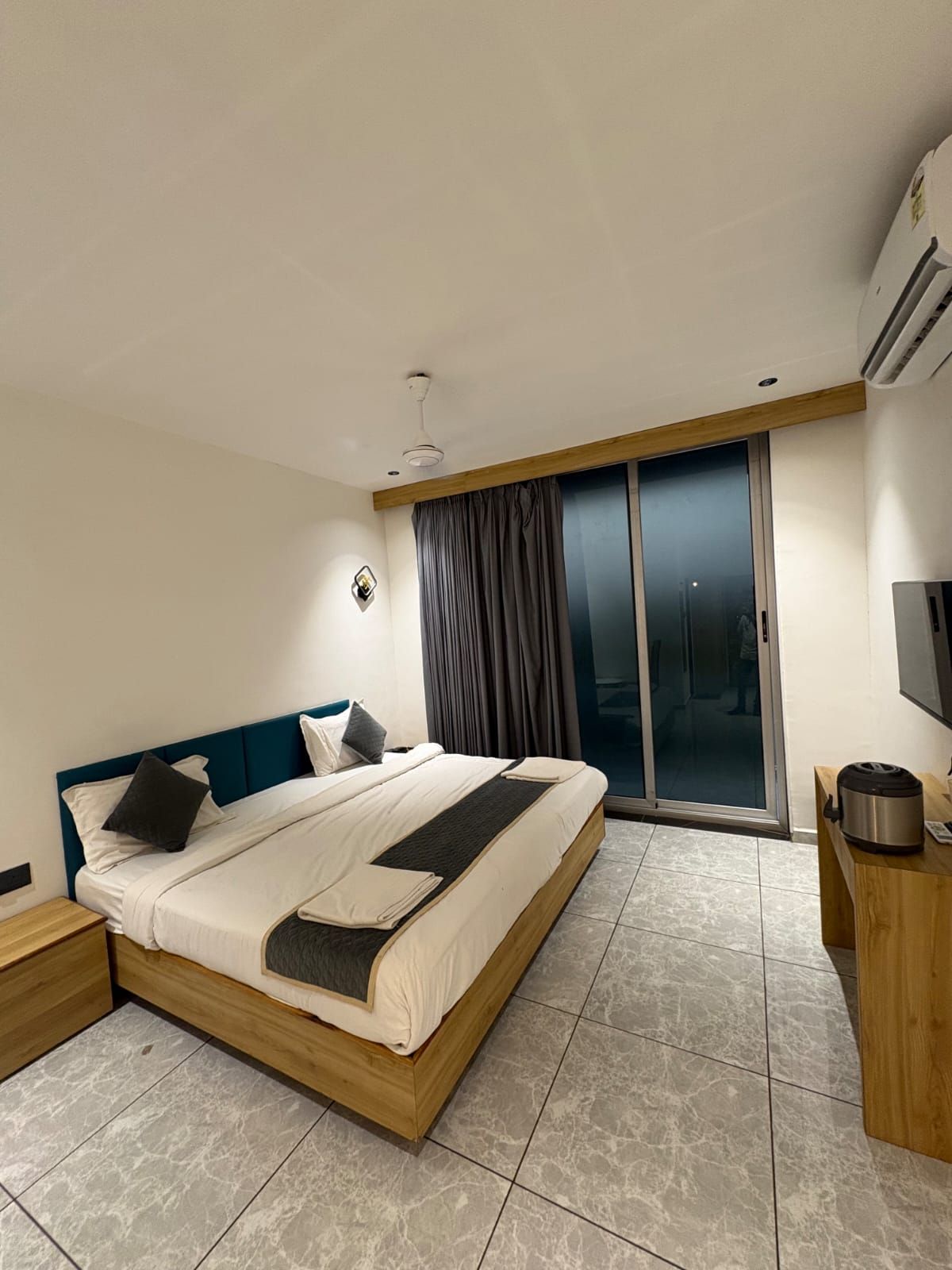 A modern hotel room with a large bed, a wooden headboard, a flat-screen TV, and a sliding glass door leading to a balcony.
