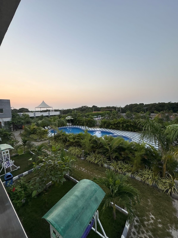 A pool area with a checkered pattern around the edge, surrounded by greenery and a white structure in the background.