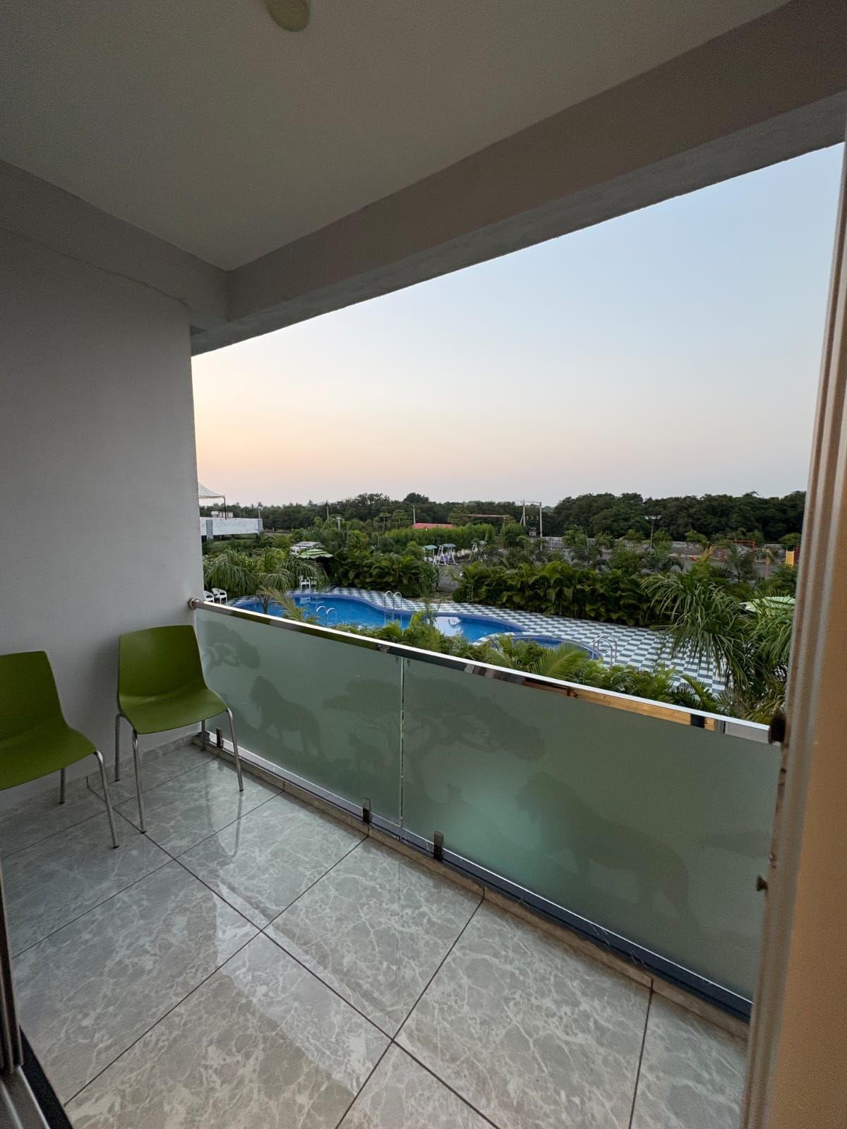 Un balcón con dos sillas verdes, barandilla de cristal y vistas a una piscina y follaje.