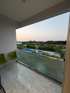 A balcony with two green chairs, a glass balustrade, and a view of a swimming pool and greenery. - Avatara Resort Sasan Gir (Talala)