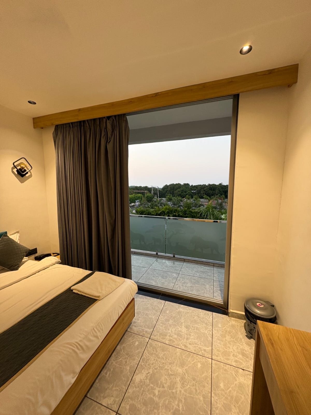 A hotel room with a bed, a small bedside table, a nightstand, a window with curtains, and a view of the outdoors.