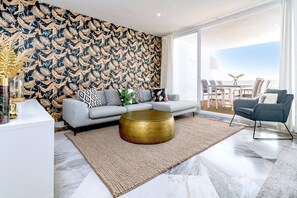 A modern living room with a patterned wallpaper, a grey sofa, a round gold coffee table, and a view through large windows. - ELD2-Luxury 3 Bedroom Penthouse in Nueva Andalucia (Marbella)