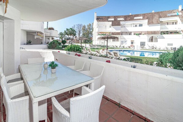 Una terraza con mesa de vidrio y sillas de mimbre, con vistas a una piscina y un edificio con balcones.
