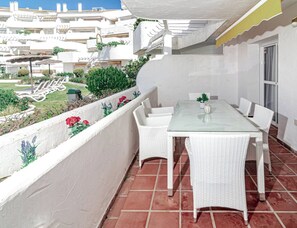 Un balcón con una mesa de vidrio y sillas de mimbre blanco, con vista a una zona residencial con tejas de barro.
