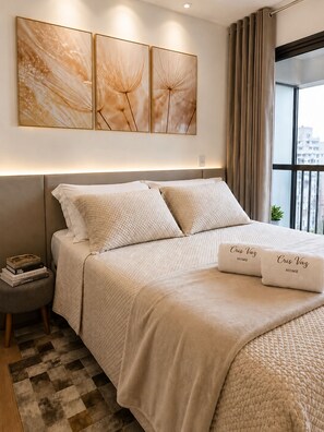 Ein ordentlich bezogenes Bett mit einer gesteppten Decke, ein Nachttisch mit Büchern und Blick auf die Stadt durch ein Fenster.