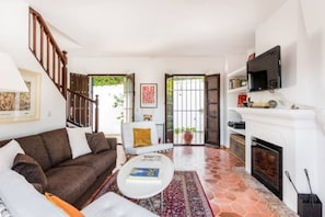 A living room with a brown sofa, white armchair, and a coffee table. There is a fireplace, a TV mounted on the wall, and a staircase leading to another level. - EN- Cozy Andalusian style townhouse in Marbella (Marbella)