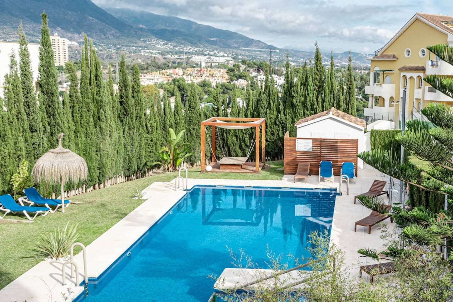 A swimming pool with a wooden swing set, lounge chairs, and a view of a cityscape with mountains in the background.