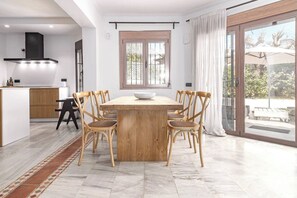 Eine moderne Küche mit einer zentralen Kochinsel, ein Essbereich mit Holztisch und -stühlen sowie eine Glastür, die auf eine Außenpatio führt.