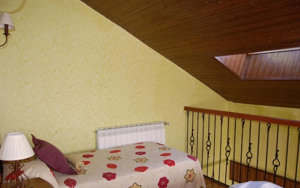 Un dormitorio con techo inclinado, una cama con ropa de cama floral, un radiador, una mesita de noche con una lámpara y una ventana con marco de madera.