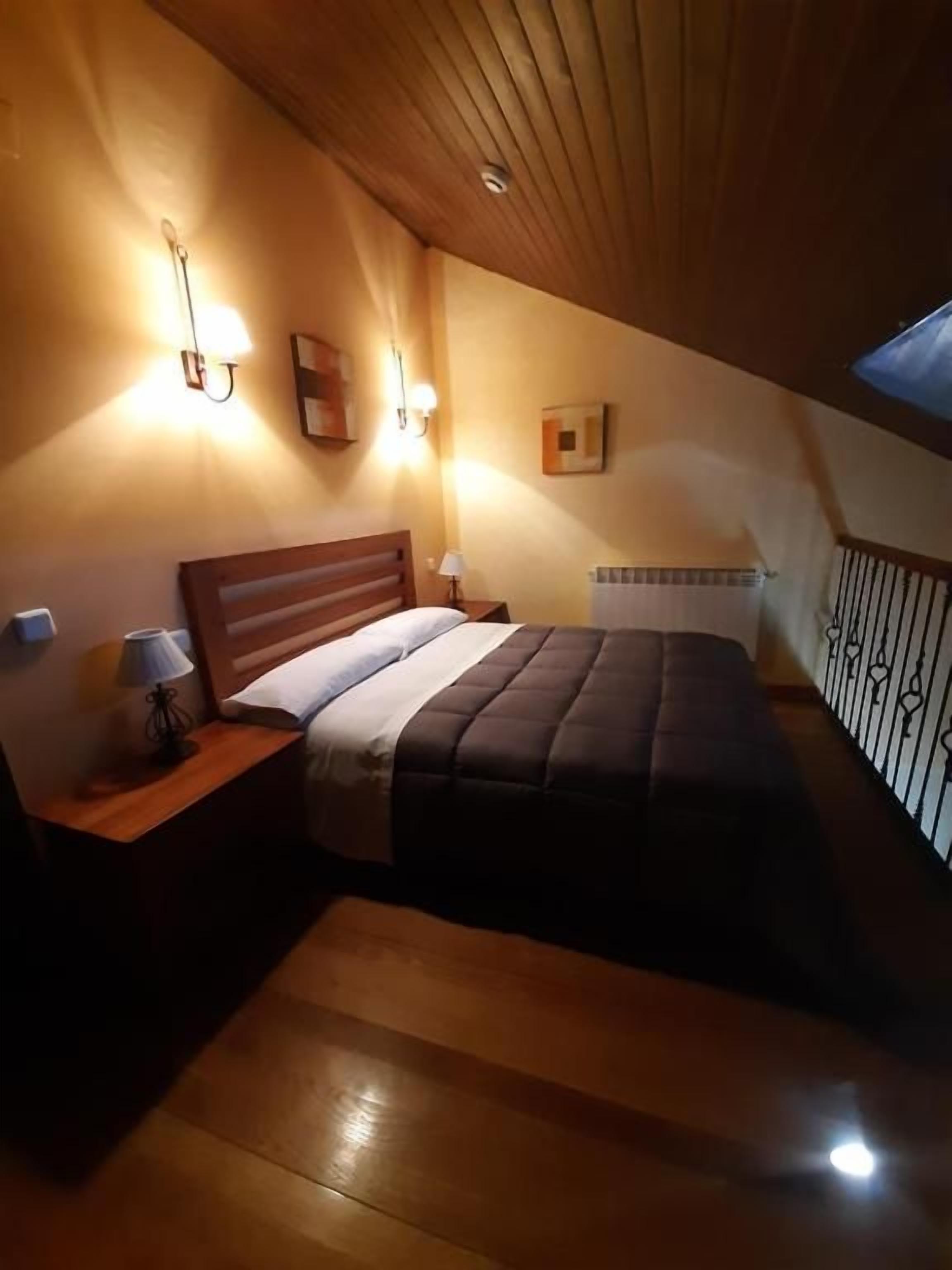 Un dormitorio con techo de madera, una cama con cubrecama oscuro, lámparas de noche y una mesita de noche.