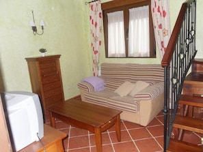 Una sala de estar con un sofá, una mesa de madera, un televisor, una escalera y un ventanal con cortinas.