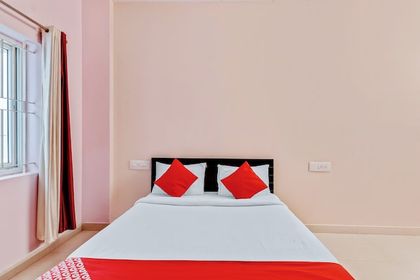A single bed with a white mattress and two red pillows, a window with white blinds, and a wall switch.