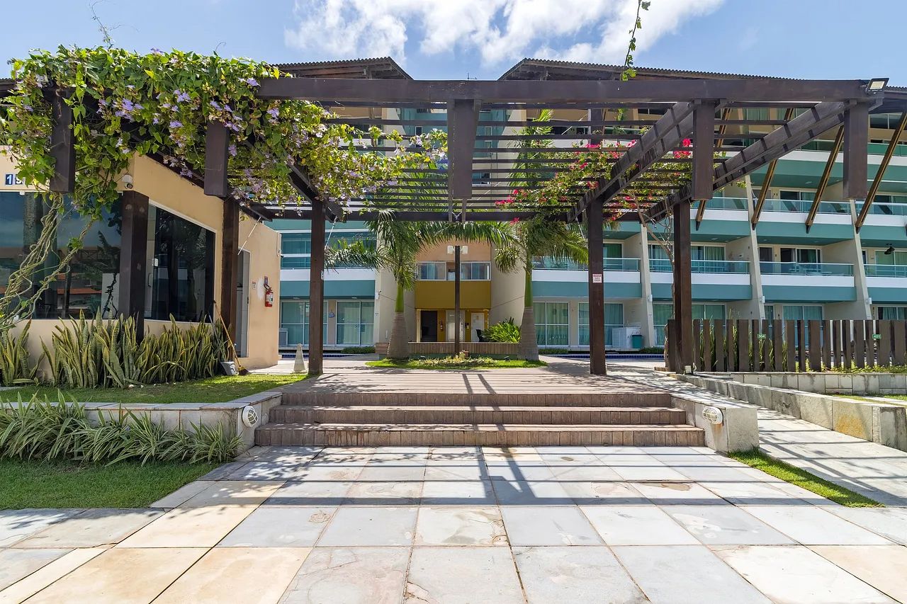 Um edifício moderno com entrada coberta, degraus que levam à entrada e área de jardim com plantas e uma pérgula.