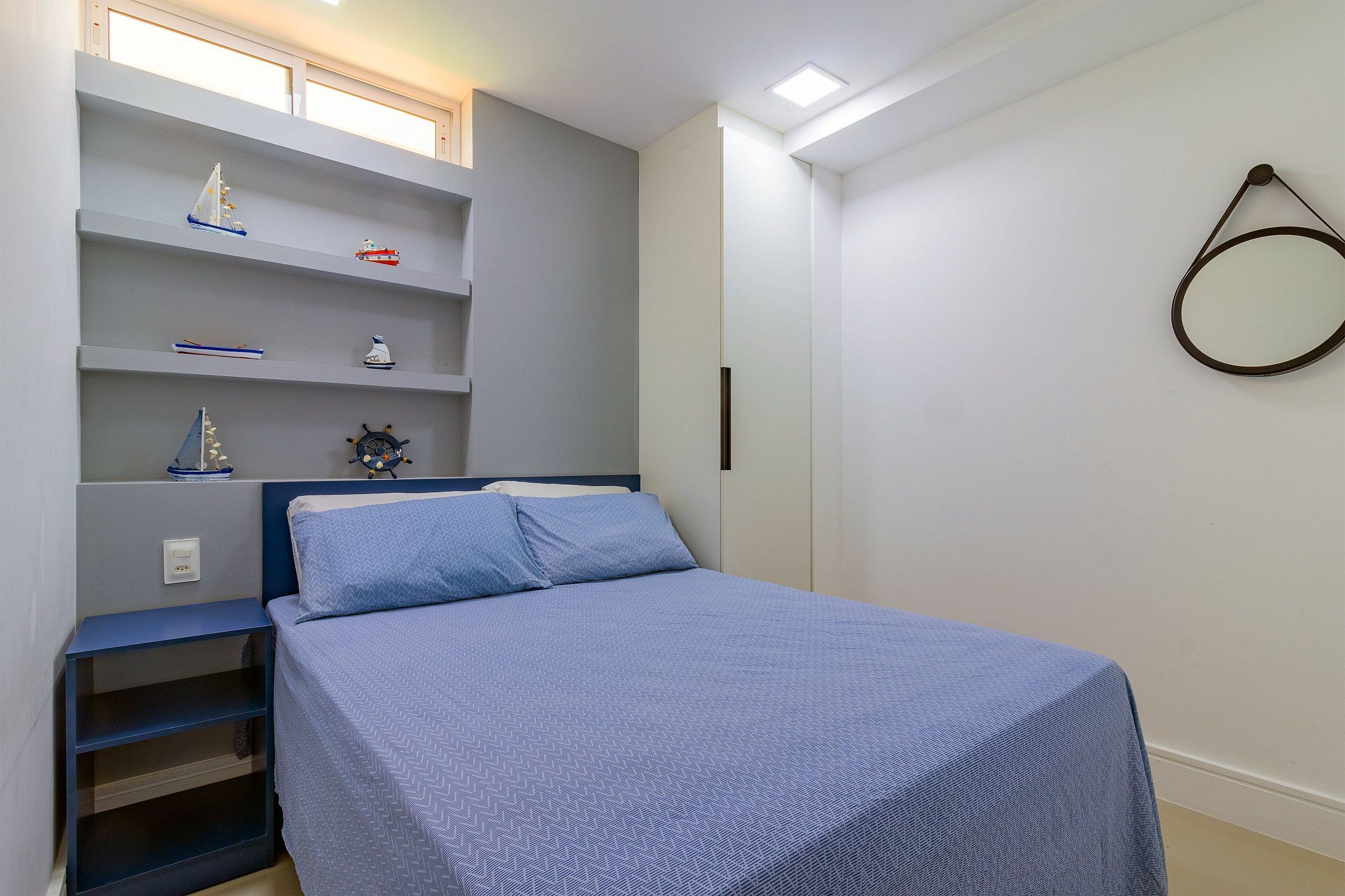 A bedroom with a bed, a shelf with decorative items, a wall-mounted mirror, and a window with natural light.