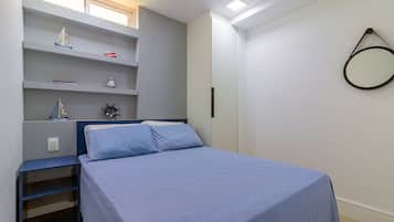 A bedroom with a bed, a shelf with decorative items, a wall-mounted mirror, and a window with natural light.