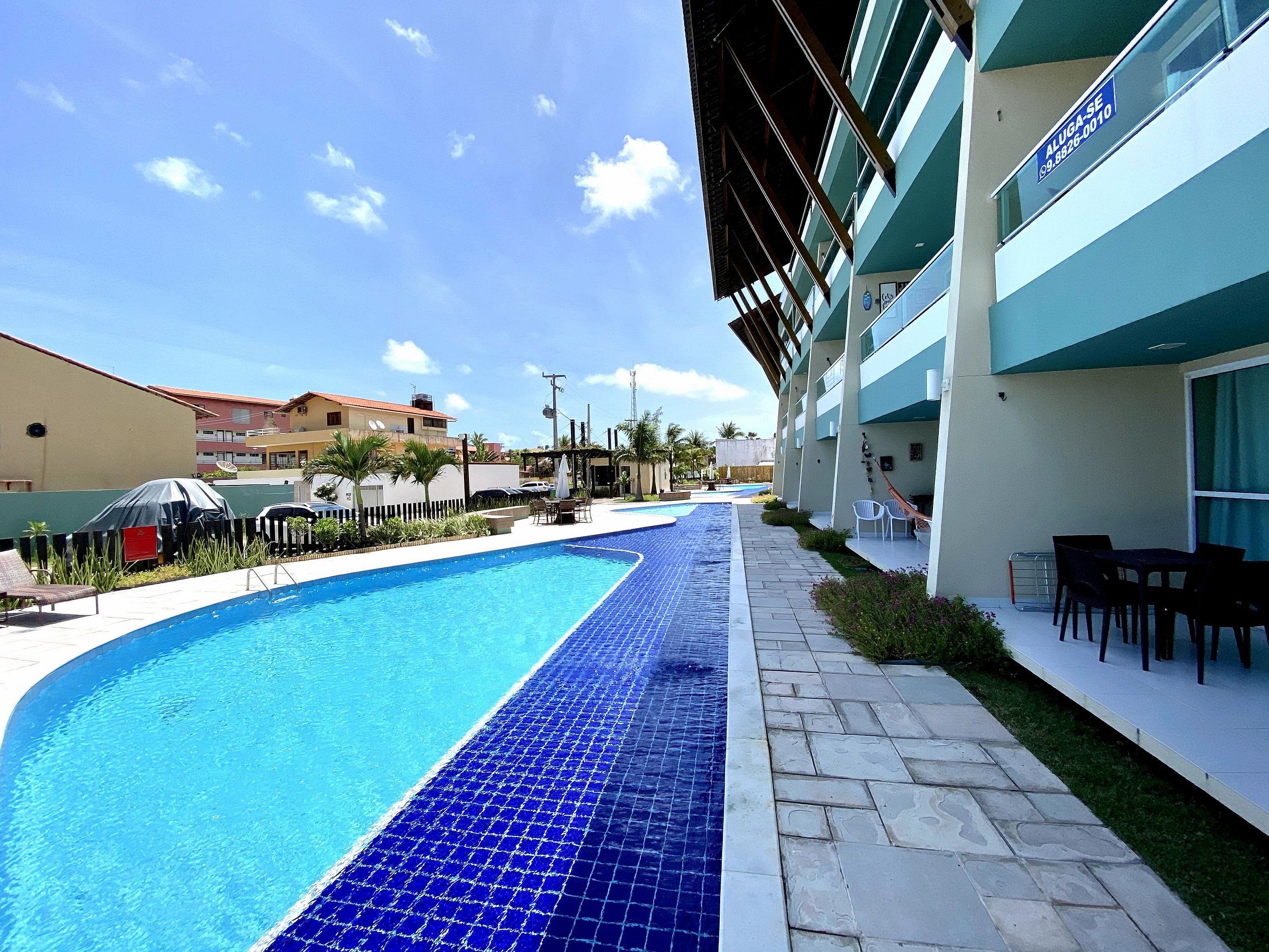 Piscina com borda em azulejo, ao lado de um edifício com varanda e área de estar externa.