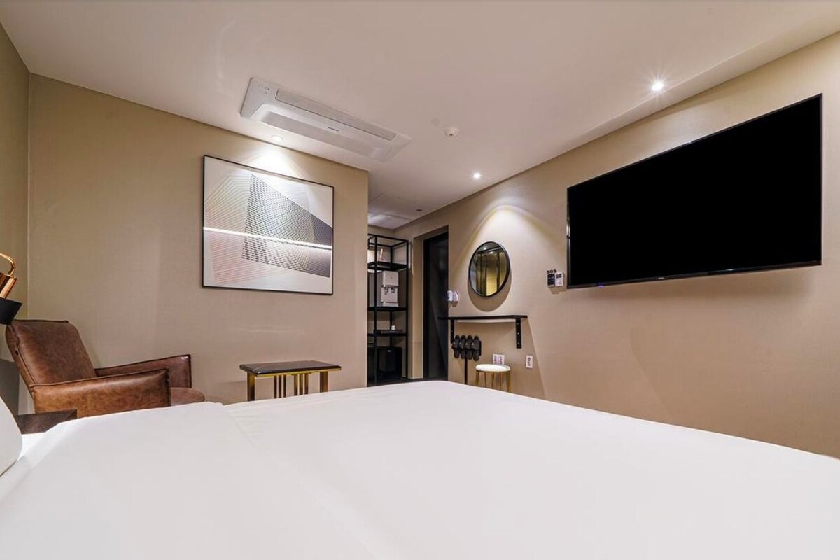 A hotel room with a large bed, a flat-screen TV mounted on the wall, a seating area with a chair and a small table, and a bookshelf.