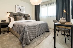 A modern bedroom with a large bed, a bedside table, a desk with a chair, and a large window with curtains. - IN33K- Stunning city apartment close to beaches (Estepona)
