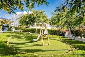 A playground with a slide and swings, surrounded by well-maintained grass and trees, with buildings in the background. - GH-Modern 2 bed apartment with Pool in Estepona (Estepona)