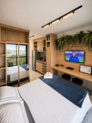 Ein modernes Hotelzimmer mit Bett, Schreibtisch mit Fernseher, Balkon mit Blick auf die Stadt und einer Küchenzeile.