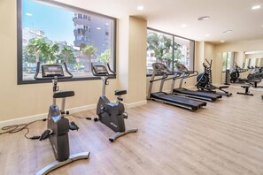 Een fitnessruimte met cardiotoestellen, waaronder loopbanden en hometrainers, en grote ramen met uitzicht op gebouwen en palmbomen.