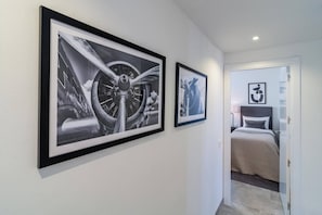 Een gang met twee zwart-wit foto's in lijsten aan de muur, die leidt naar een slaapkamer met een bed en een nachtkastje.
