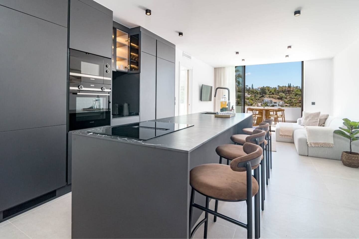 A modern kitchen with a central island and bar stools, a dining area with a table and chairs, and a view of a hillside.