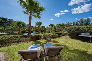 A patio with two cushioned chairs, a grill, and a view of a lake and palm trees. - LAlq13 Stunning Frontline Golf 3 bed apt (Benahavís)