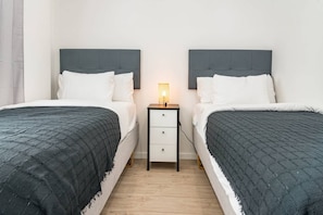 Een tweepersoonskamer met twee bedden, elk met een grijze hoofdbord, witte beddengoed en een grijs gebogen dekbed. Daartussen staat een klein nachtkastje met een lamp.