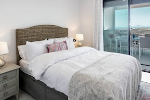 A hotel room with a bed, a nightstand, a lamp, a patterned throw, and a view of the city through a large window. - Luxury Resort Apt with Sea View & Pools - LAE23i (Estepona)