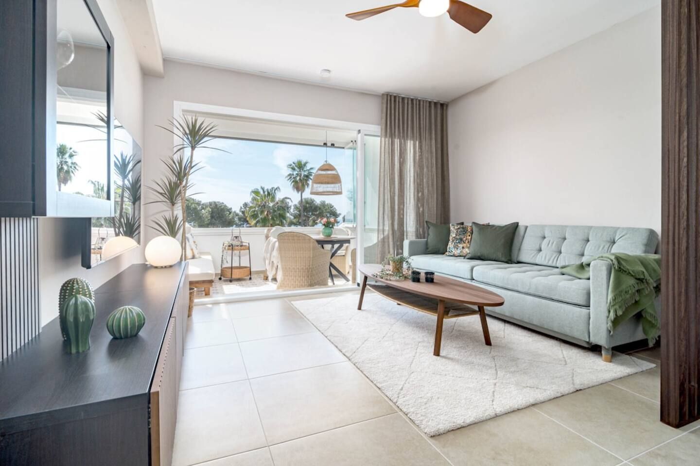 A modern living room with a sofa, coffee table, and a TV on a stand. There is a balcony with plants and a view of palm trees.