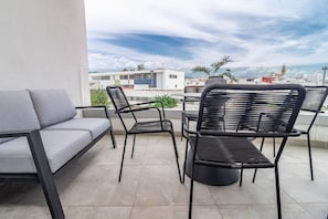Eine Terrasse mit einer Couch und zwei Stühlen, im Hintergrund eine Ansicht von Gebäuden und einem Gewässer.