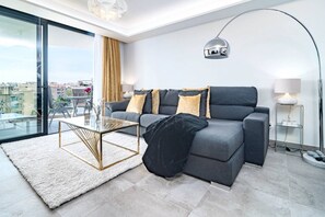 A modern living room with a grey sofa, a glass coffee table, a black throw blanket, a floor lamp, and a view of the city through large windows. - INf45P- modern 3 bed city apartment next to beach (Estepona)