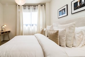 A hotel room with a large bed, a bedside table with a lamp, a window with sheer curtains, and framed pictures on the wall. - INf45P- modern 3 bed city apartment next to beach (Estepona)