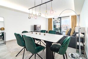 A modern living room with a dining area, green velvet chairs, a white table, and a city view through large windows. - INf45P- modern 3 bed city apartment next to beach (Estepona)