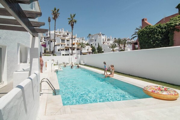 A swimming pool with clear blue water, surrounded by white buildings and palm trees. - IVY - Scandinavian Apartment in Nueva Andalucia (Marbella)