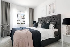 A modern bedroom with a tufted headboard, a large bed with multiple pillows, a bedside table with a lamp, and a view of the outside through a window. - IVY - Scandinavian Apartment in Nueva Andalucia (Marbella)